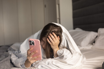 young woman is sitting on bed wrapped in blanket appearing stressed as she holds a smartphone. Her expression shows concern or frustration.indicating that she is absorbed in something on the device