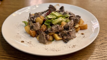 Warm salad with chicken liver in cream sauce with fried potatoes, mushrooms and mixed green salad in a white porcelain plate on a brown wooden table in an elite restaurant. Close-up