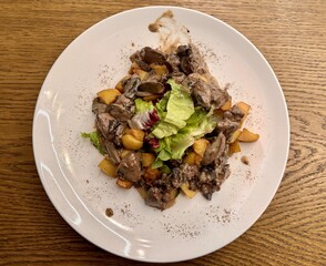Warm salad with chicken liver in cream sauce with fried potatoes, mushrooms and mixed green salad in a white porcelain plate on a brown wooden table in an elite restaurant. Close-up