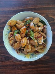 Stir-fried clams with chili paste and basil leaves in a white bowl on a wooden table.