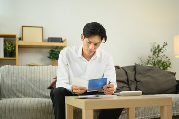Fototapeta premium Serious asian man reviewing bank statements and calculating expenses in a living room