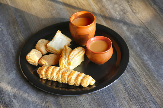 Tabletop shot of tea cups with khari (salty puff pastry), cream roll, and toasted bread or Jeera Butter.