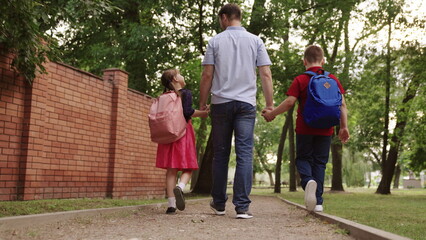 Fototapeta premium happy family. father holding hands children school. little kids school bags go school holding dad hand. concept happy family outdoors. schoolchildren with parent view from back. children education