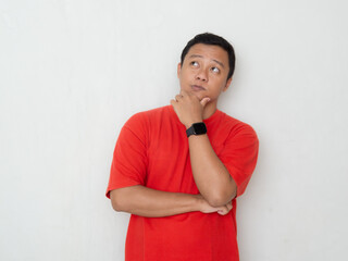 Young Asian man in red shirt stands thoughtfully with his hand on the chin and arms crossed over...