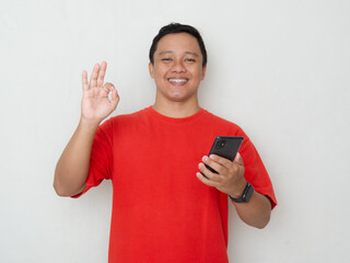 Young Asian man in red shirt giving an OK hand gesture while holding his phone over white...