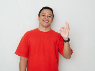 Smiling young Asian man in red shirt giving an OK hand gesture over white background. The image...