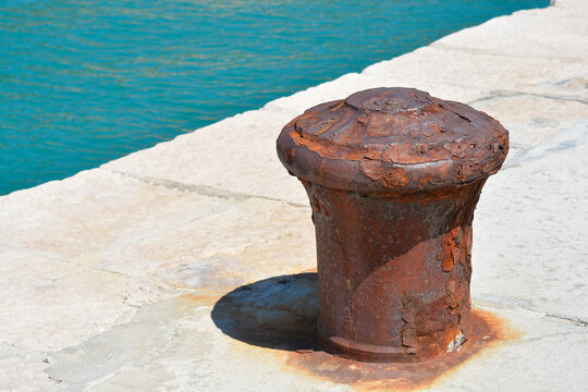 A rusty mooring bollard on a stone dock next to a turquoise sea