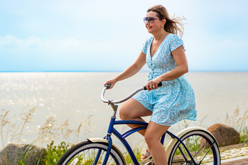 Active summer holidays. Mature woman wearing dress riding blue cruiser bike along watershore in...