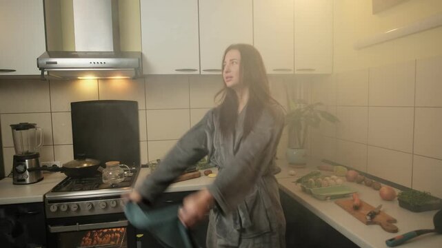 A housewife in a soft gray bathrobe frantically waves a towel to disperse smoke from burnt food in a cozy kitchen filled with various ingredients.