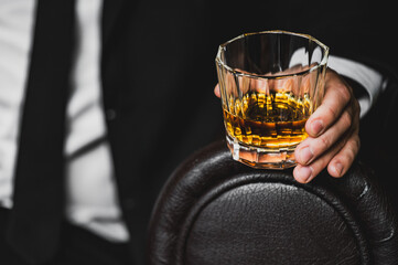 A close-up of a hand holding a crystal glass filled with whiskey. The background features a dark,...