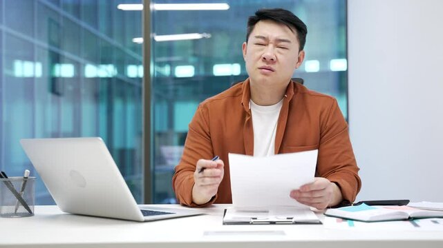 Overworked upset asian businessman is tired of doing paperwork sitting at workplace at desk in modern business office. Lazy exhausted man worker holds a document, procrastinates, gets distracted