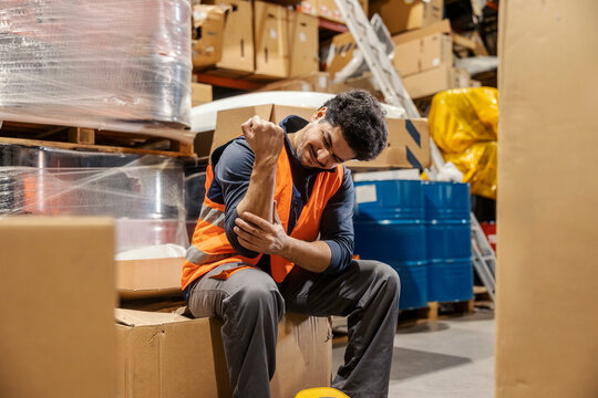 Interracial storage worker with injured arm sitting at warehouse surrounded by boxes and shipment.