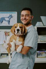 Veterinarian smiling while holding a beagle puppy in well-equipped veterinary clinic featuring medical posters and workspace in background
