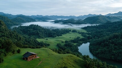 Obraz premium Wooden cabin sits on a green grassy hill overlooking a winding river and misty mountain valley at dawn.