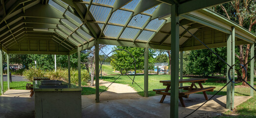 Panoramic view of a public park shelter featuring a covered BBQ area with picnic tables and translucent roofing. The shelter opens to a well-maintained outdoor recreational space. Victoria, Australia