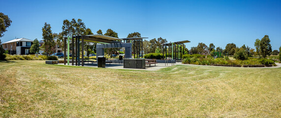 Panoramic view of a modern suburban park with a shaded picnic area featuring barbecue grills and...