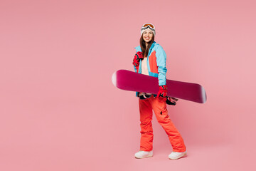Full body side view young smiling woman wear warm blue padded jacket ski goggles mask spend weekend winter season in mountains hold snowboard walk go isolated on plain pink background. Hobby concept.