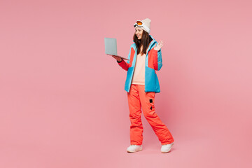 Full body skier young woman wear warm blue padded jacket ski goggles mask spend weekend winter season in mountains hold use work on laptop pc computer isolated on plain pink background. Hobby concept. © ViDi Studio