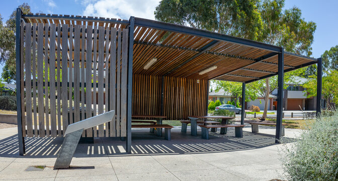 A modern outdoor public shelter or pavilion with a contemporary architectural design and vertical wooden slats. A stylish community rest area with picnic table and built-in seating in a suburban park.