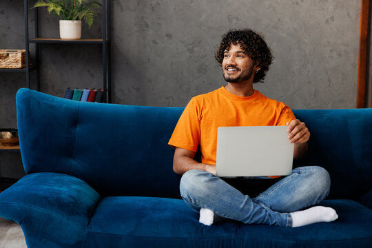 Young IT Indian man wear orange casual clothes sit on blue sofa couch hold use work on laptop pc computer stay home hotel flat rest relax spend free spare time in living room indoor. Lounge concept.