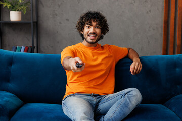 Young smiling happy Indian man he wear orange casual clothes sits on blue sofa couch stay at home...