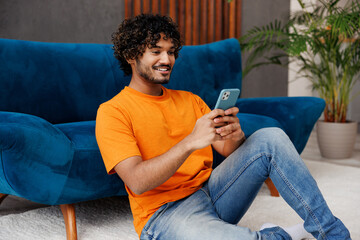 Side view young Indian man wear orange casual clothes sit near blue sofa couch hold use mobile cell...