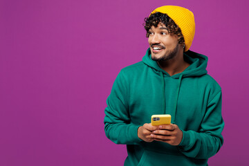 Young happy Indian man wear green hoody yellow hat casual clothes hold in hand use mobile cell phone look aside on area mock up isolated on plain purple background studio portrait. Lifestyle concept.