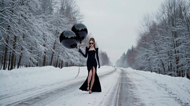 Elegant fashion model walking confidently on snowy road, wearing sleek black dress and gloves, holding two black balloons against wintery forest landscape