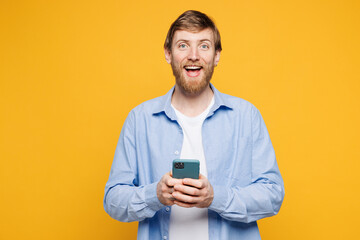 Young shocked surprised happy Caucasian man wear blue shirt t-shirt casual clothes hold in hand use mobile cell phone look camera isolated on plain yellow background studio portrait Lifestyle concept