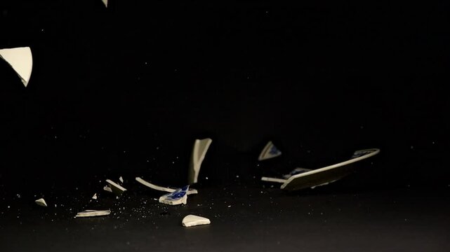 A vibrant hand-painted plate crashes to the ground, with shards scattering in slow motion. The black matte backdrop highlights the dramatic impact.
