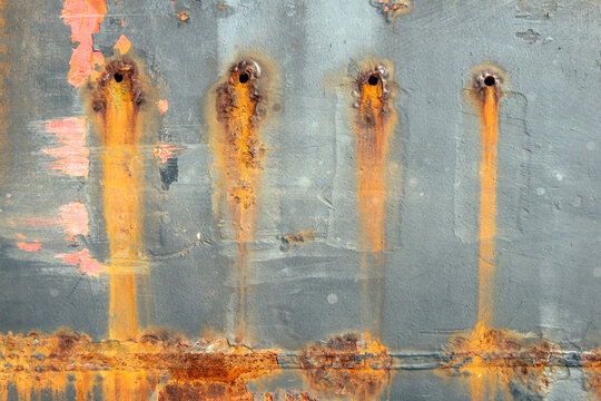 Closeup of a weathered ship hull with rust. There are four small holes in the ship's side. Here and there the red lead is visible.