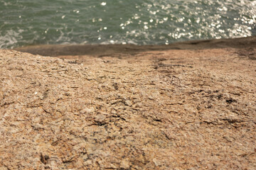 Close-up of a textured rock surface near the water. The rock looks uneven and weathered, with barely noticeable color transitions. There is a shimmering ocean in the background.