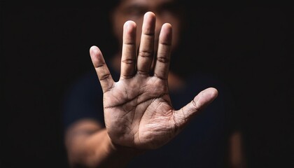 human hand in a stop gesture, palm facing outward with fingers extended, representing warning, caution, and refusal, with dramatic lighting and a blurred figure in the background
