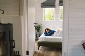 A woman lies comfortably on her back in bed, wearing a blue shirt, smiling as she stretches. Sunlight gently illuminates her peaceful expression, enhancing a serene morning atmosphere.