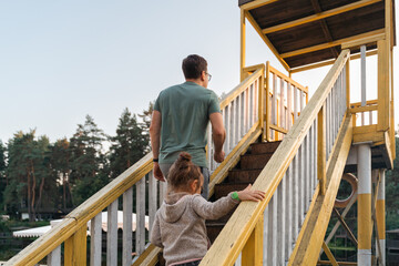 A father leads his daughter up a set of yellow wooden stairs surrounded by trees, exploring the...