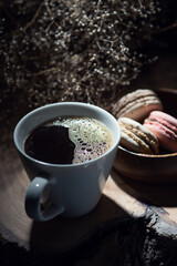 A white cup filled with black coffee sits on a wooden surface, with a bowl of assorted macarons beside it. The background features dried flowers, creating a cozy and rustic atmosphere.