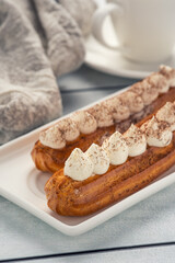 Two eclairs topped with whipped cream and cocoa powder on a rectangular white plate, with a blurred background of a white cup and saucer.