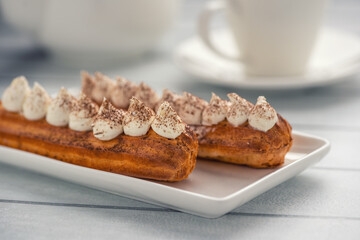 Two eclairs topped with whipped cream and cocoa powder on a rectangular white plate, with a blurred background of a white cup and saucer.
