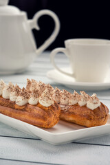 Two eclairs topped with whipped cream and cocoa powder on a rectangular white plate, with a blurred background of a white cup and saucer.