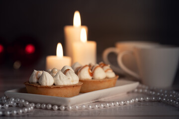 Two tarts topped with whipped cream and caramel drizzle on a white plate, surrounded by white candles, two white cups, and pearl necklaces on a wooden surface