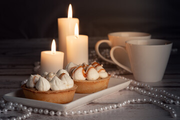 Two tarts topped with whipped cream and caramel drizzle on a white plate, surrounded by white candles, two white cups, and pearl necklaces on a wooden surface