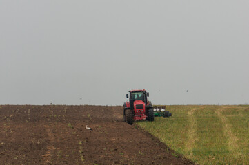 Obraz premium A red tractor is plowing a field, creating a clear division between the tilled soil on the left and the unplowed grass on the right.