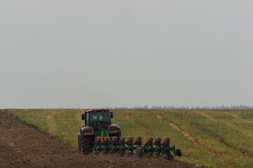 A red tractor is plowing a field, creating a clear division between the tilled soil on the left and the unplowed grass on the right.