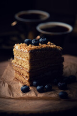 A slice of layered cake topped with blueberries sits on parchment paper, with two cups of coffee in the background, creating a cozy and inviting scene.