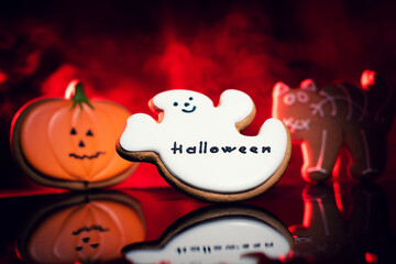 Three Halloween-themed cookies: a smiling ghost with "Halloween" written on it, a pumpkin with a jack-o'-lantern face, and a cat with a spider web design, against a red smoky background