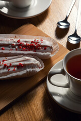 Two eclairs with white icing and red sprinkles on a wooden board, accompanied by a cup of tea and two spoons on a wooden table.