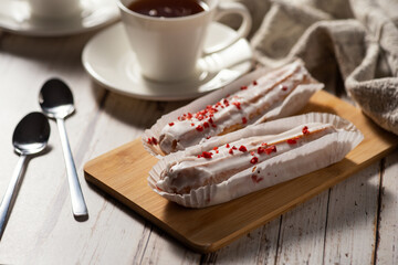 Two éclairs with white icing and red sprinkles on a wooden board, accompanied by a cup of tea and two spoons on a rustic wooden table.