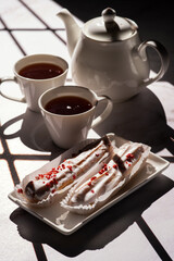 Two cups of tea, a white teapot, and two eclairs with white icing and red sprinkles on a white plate are arranged on a table with a grid shadow pattern.