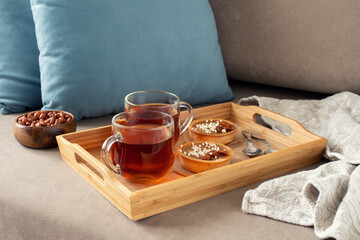 Two cups of tea on a saucer with two small tarts topped with white sprinkles, placed on a beige couch with blue pillows in the background
