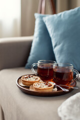 Two cups of tea on a saucer with two small tarts topped with white sprinkles, placed on a beige couch with blue pillows in the background